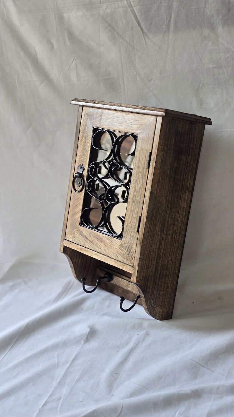 Op de afbeelding: Een houten wandkast met een donkere afwerking. De kast heeft een decoratief metalen ontwerp achter de glazen deur en twee zwarte haken eronder. Ontworpen voor opslag en organisatie.