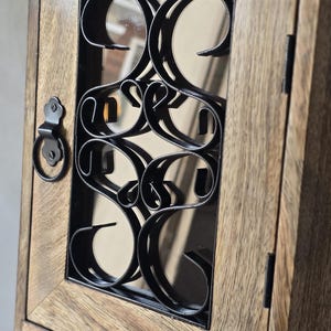 Op de afbeelding: Houten wandkast met een decoratief zwart metalen ontwerp op de glazen deur. De kast heeft een lichtbruine afwerking en een kleine handgreep. De deur is gesloten en de kast lijkt leeg te zijn.