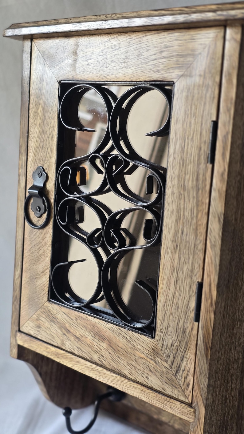 Op de afbeelding: Houten wandkast met een decoratief zwart metalen ontwerp op de glazen deur. De kast heeft een lichtbruine afwerking en een kleine handgreep. De deur is gesloten en de kast lijkt leeg te zijn.