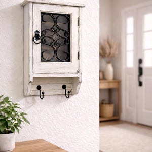 Op de afbeelding: Een witte, aan de muur gemonteerde kast met een verweerde afwerking. Het heeft een glazen deur met een decoratief zwart metalen ontwerp en twee zwarte haken eronder. De kast is gemonteerd op een witte, getextureerde muur.