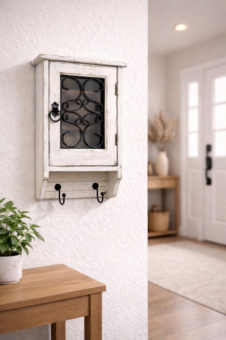 Op de afbeelding: Een witte, aan de muur gemonteerde kast met een verweerde afwerking. Het heeft een glazen deur met een decoratief zwart metalen ontwerp en twee zwarte haken eronder. De kast is gemonteerd op een witte, getextureerde muur.