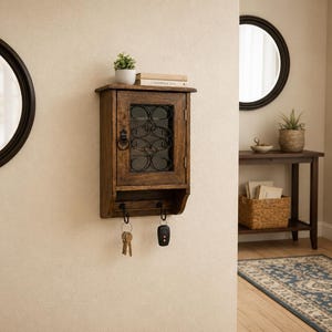 Op de afbeelding: Een houten sleutelhouder voor aan de muur met een glazen deur en decoratief metaalwerk. De sleutelhouder heeft een klein plankje met een plant en boeken. Hieronder haken met sleutels en een autosleutel. Twee ronde zwarte spiegels zijn zichtbaar.