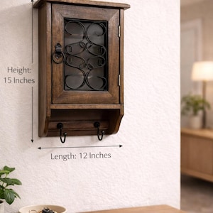 Op de afbeelding: Een houten wandkast met een glazen deur en decoratieve metalen details. De kast is 38 cm hoog en 30 cm lang en heeft twee haken eronder. Het is aan een witte muur bevestigd, geschikt voor opslag of weergave.