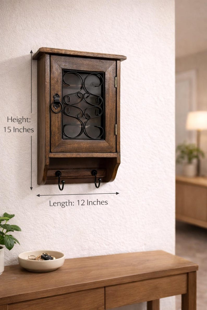 Op de afbeelding: Een houten wandkast met een glazen deur en decoratieve metalen details. De kast is 38 cm hoog en 30 cm lang en heeft twee haken eronder. Het is aan een witte muur bevestigd, geschikt voor opslag of weergave.