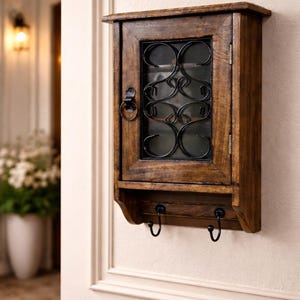 Op de afbeelding: Een houten sleutelkastje met een donkere afwerking, met een glazen deur met decoratief zwart metalen krulwerk. Onder het kastje bevinden zich twee zwarte haken. Het kastje is aan een lichtgekleurde muur bevestigd.