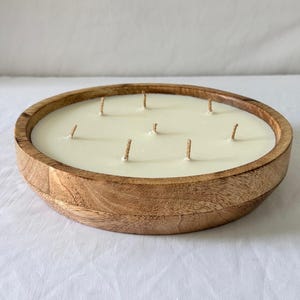 May include: A large, round candle in a wooden bowl. The candle is ivory-colored with multiple wicks. The wooden bowl has a natural wood grain finish. The candle is likely scented and designed for home decor.