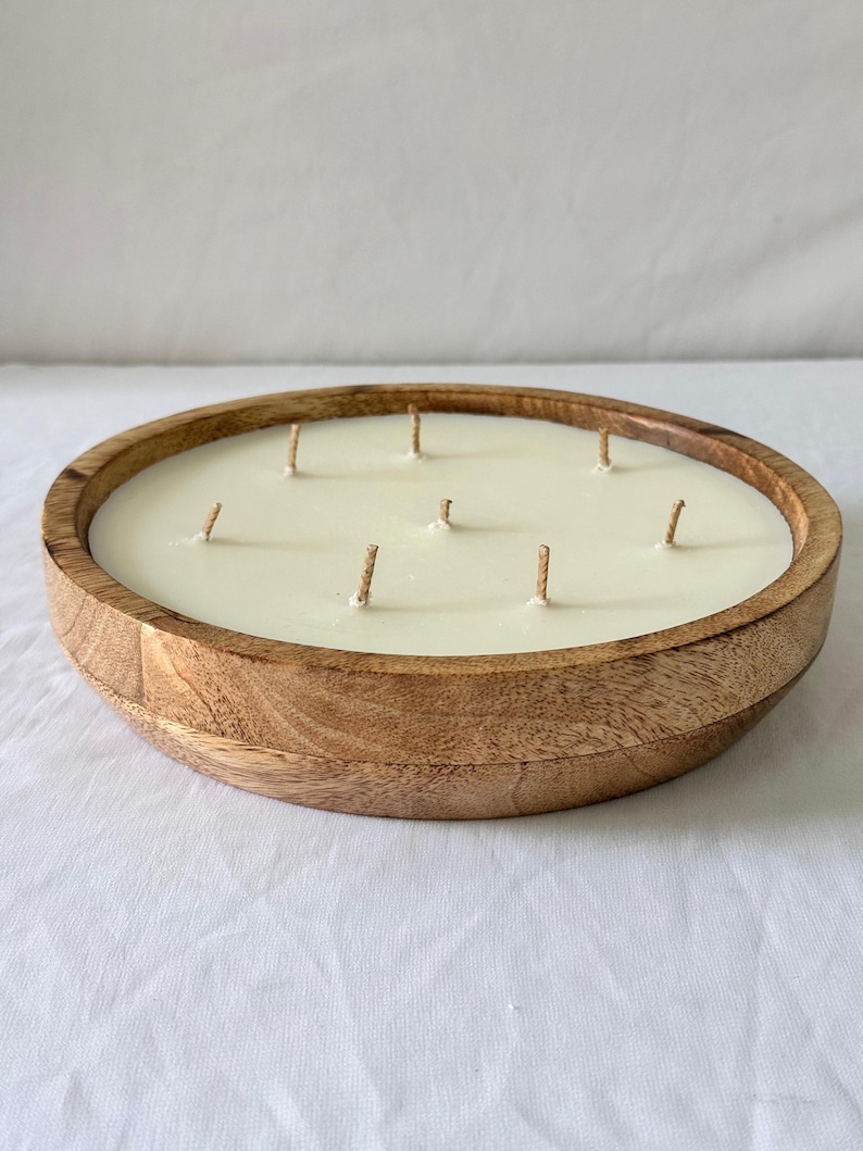 May include: A large, round candle in a wooden bowl. The candle is ivory-colored with multiple wicks. The wooden bowl has a natural wood grain finish. The candle is likely scented and designed for home decor.