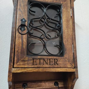 Op de afbeelding: Een houten sleutelhouder voor wandmontage met een donkerbruine afwerking. De kastdeur heeft een decoratief zwart metalen ontwerp en het woord "ETNER" is op de voorkant gegraveerd. Onder de kast bevinden zich twee zwarte haken.