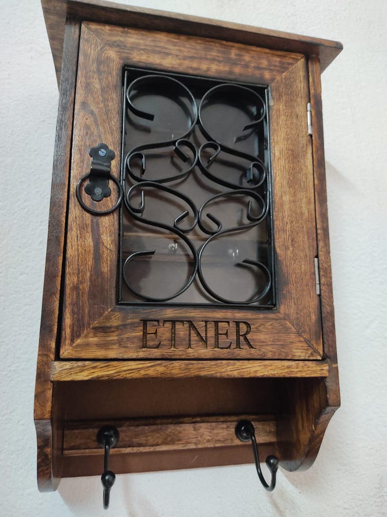 Op de afbeelding: Een houten sleutelhouder voor wandmontage met een donkerbruine afwerking. De kastdeur heeft een decoratief zwart metalen ontwerp en het woord "ETNER" is op de voorkant gegraveerd. Onder de kast bevinden zich twee zwarte haken.