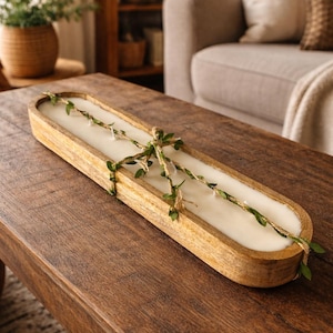 Op de afbeelding: Een lange, houten kandelaar met een crèmewitte kaars erin. De kaars is versierd met een slinger van touw en groen. De houder staat op een donkere houten tafel, wat een rustieke, boerderijachtige esthetiek creëert.