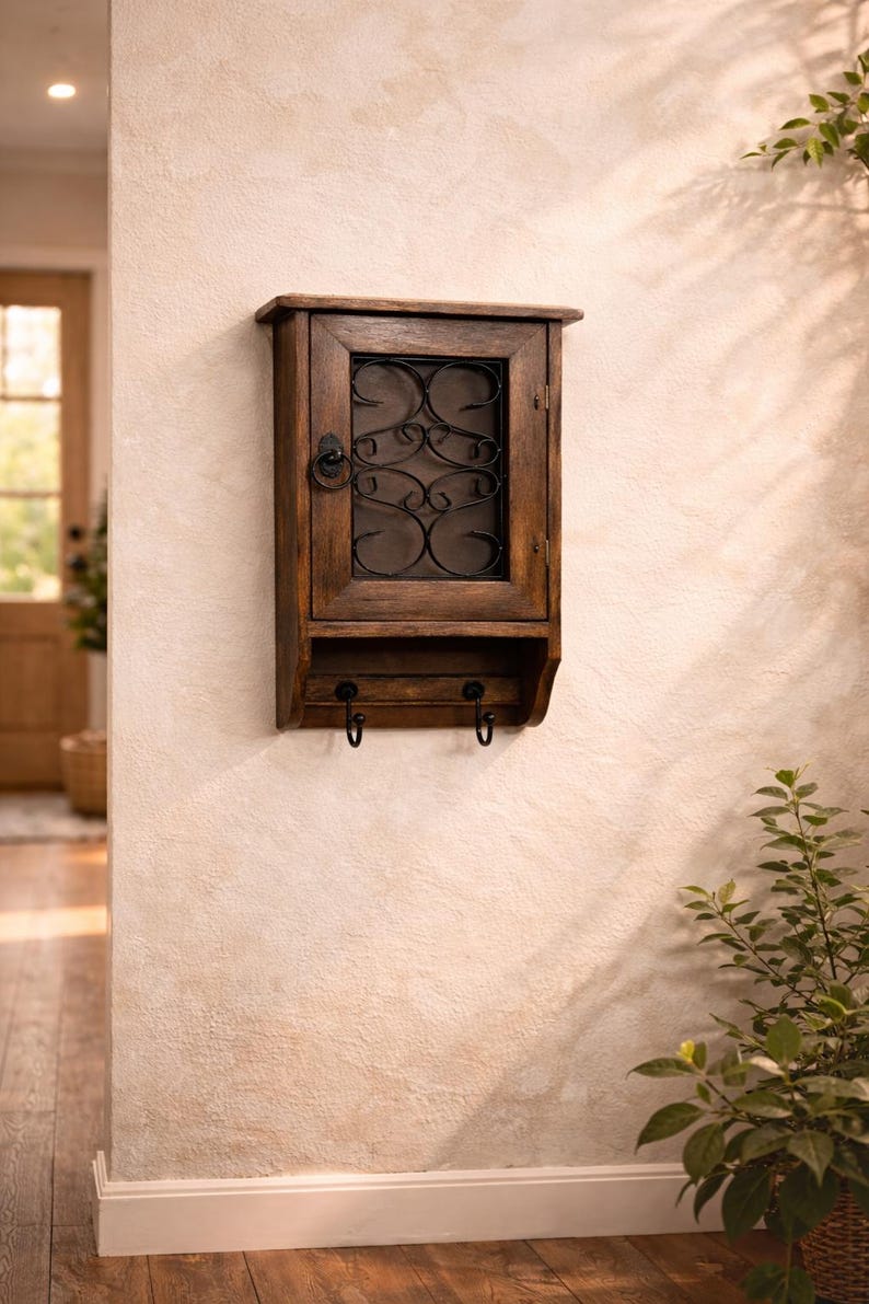 Op de afbeelding: Een donkerhouten wandkast met een glazen deur en decoratief metalen krulwerk. Onder de kast bevinden zich twee haken. De kast is bevestigd aan een getextureerde beige muur.