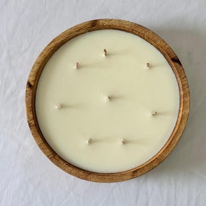 May include: A large, round candle with multiple wicks sits in a wooden bowl. The candle wax is a creamy off-white color. The wooden bowl has a natural wood grain finish.