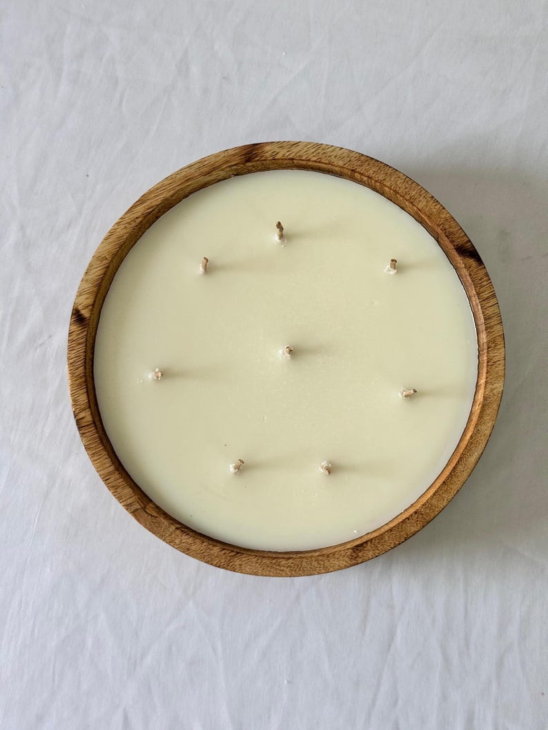 May include: A large, round candle with multiple wicks sits in a wooden bowl. The candle wax is a creamy off-white color. The wooden bowl has a natural wood grain finish.