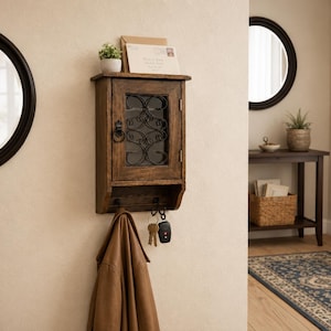 Op de afbeelding: Een rustieke houten wandkast met een glazen deur en decoratief ijzerwerk. Daaronder een plank met sleutels en een bruine leren jas. Een kleine plant en post liggen bovenop. Een ronde spiegel staat links.