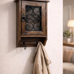 Op de afbeelding: Een houten wandkast met een donkere afwerking, met een glazen deur met een decoratief metalen overlay. Onder de kast bevinden zich drie haken, met een beige jas die aan &eacute;&eacute;n ervan hangt. De kast is gemonteerd op een witte muur. Ongeveer 30 cm breed.