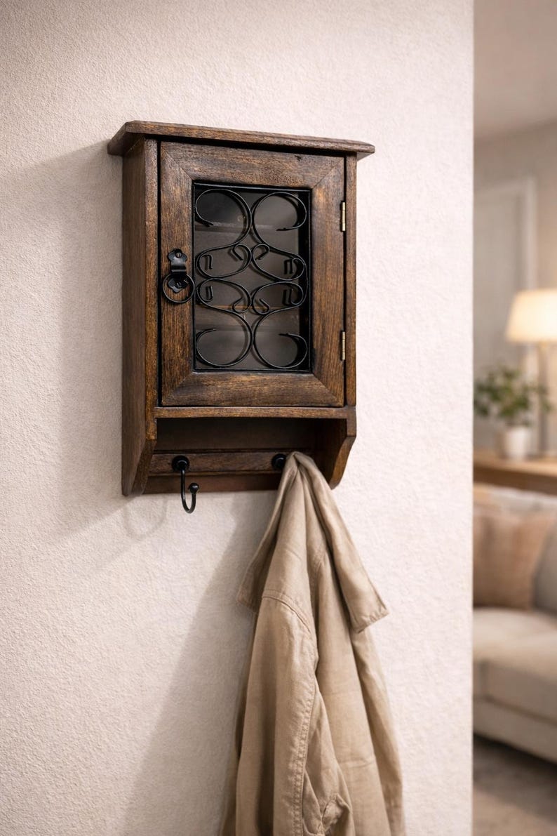 Op de afbeelding: Een houten wandkast met een donkere afwerking, met een glazen deur met een decoratief metalen overlay. Onder de kast bevinden zich drie haken, met een beige jas die aan &eacute;&eacute;n ervan hangt. De kast is gemonteerd op een witte muur. Ongeveer 30 cm breed.