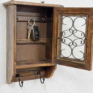Op de afbeelding: Een donkerbruine houten sleutelhouder. Het heeft een klein plankje, een deur met een decoratief metalen ontwerp en sleutels die erin hangen. Onder het plankje zitten twee zwarte haken. De sleutelhouder is aan een witte muur bevestigd.