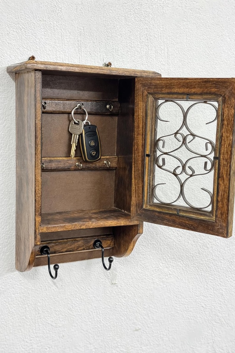 Op de afbeelding: Een donkerbruine houten sleutelhouder. Het heeft een klein plankje, een deur met een decoratief metalen ontwerp en sleutels die erin hangen. Onder het plankje zitten twee zwarte haken. De sleutelhouder is aan een witte muur bevestigd.