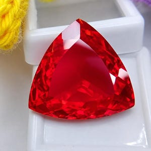Könnte beinhalten: Ein leuchtend roter, transparenter Edelstein in dreieckiger Form mit zahlreichen Facetten, die das Licht reflektieren. Der Stein besticht durch einen brillanten karmesinroten Farbton und eine glatte, polierte Oberfläche mit klarem, geometrischem Design.