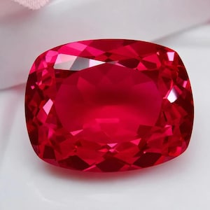 Può includere: Un grande rubino taglio a cuscino. La gemma è di un colore rosso intenso e vibrante, con molteplici sfaccettature che riflettono la luce. La forma è rettangolare con angoli arrotondati e la superficie è molto lucida.