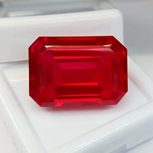 Peut inclure: Une pierre précieuse rectangulaire de taille émeraude d'un rouge profond et saturé. La pierre présente des bords précis à facettes multiples qui créent une surface claire et réfléchissante, mettant en valeur sa transparence et sa silhouette géométrique nette.