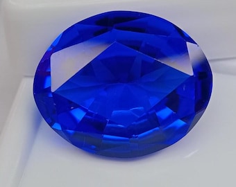 May include: A vibrant, oval-cut gemstone with a deep, saturated blue color. The stone features numerous triangular and trapezoidal facets that create a complex, geometric pattern across its surface, reflecting light with high clarity and brilliance.