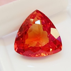 May include: A faceted, triangular gemstone with a vibrant blend of orange and red hues. The gem's cut reveals multiple facets, creating a play of light and shadow. The gemstone is set against a white background.