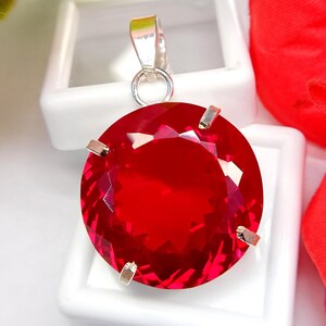 以下が含まれることがあります： 銀色の金属製の爪留め枠にセットされた、鮮やかな赤色の円形でファセットカットされた宝石のペンダント。円形の石は複数の反射面を持つブリリアントカットが施されており、上部には銀色の金属製のバチカンが取り付けられています。