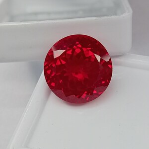 Può includere: Una pietra preziosa rotonda e sfaccettata di un colore rosso intenso e vibrante. La pietra presenta un taglio brillante con numerose sfaccettature triangolari e a forma di aquilone che riflettono la luce, creando un aspetto scintillante.