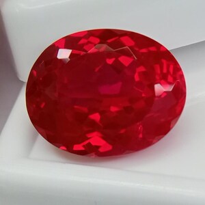 Peut inclure: Une pierre précieuse de couleur rouge vif, taillée en forme ovale avec une surface à facettes qui reflète la lumière. La pierre présente une finition polie avec de multiples facettes géométriques sur le dessus, créant un aspect brillant.