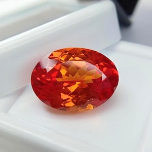 May include: An oval-shaped gemstone, displaying a rich mix of red and orange tones. The gem is cut with facets, creating a play of light. The stone is set against a white backdrop, accentuating its colour and clarity. The gem is likely a precious stone.