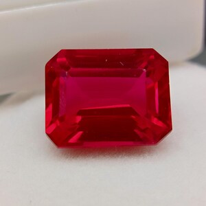 Könnte beinhalten: Ein leuchtender, rechteckig geschliffener Edelstein in einer tiefen, gesättigten roten Farbe. Der Stein weist eine facettierte Oberfläche auf, die das Licht reflektiert und ein klares, transparentes Erscheinungsbild mit scharfen Kanten erzeugt.
