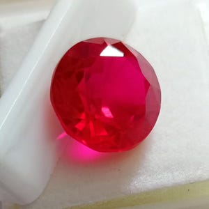 Peut inclure: Une pierre précieuse ronde à facettes d'une couleur rouge vive et saturée. La pierre présente une taille brillant avec de multiples facettes triangulaires et en forme de cerf-volant qui reflètent la lumière, créant des reflets sur sa surface.