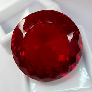May include: A round, faceted gemstone with a deep, vibrant red color. The stone features a precise cut with multiple triangular and kite-shaped facets that reflect light across its surface, creating a brilliant, sparkling appearance throughout.