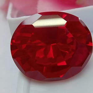 Peut inclure: Une pierre précieuse rouge de taille ovale avec de multiples facettes qui reflètent la lumière sur sa surface. La pierre présente une teinte cramoisie profonde et saturée, placée sur un fond blanc, mettant en valeur sa structure claire et polie.