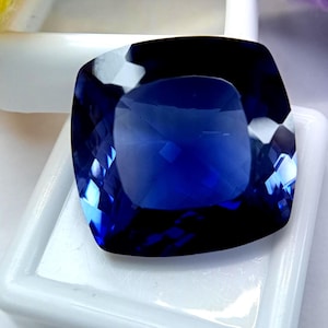 May include: A cushion-cut gemstone with a deep, saturated royal blue color and faceted surfaces that reflect light. The stone is positioned on a white square display base, showcasing its transparent quality and geometric shape with rounded corners.