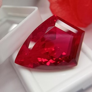 Pode incluir: Uma pedra preciosa facetada em forma de escudo com uma cor vermelha profunda e vibrante. A pedra apresenta múltiplos cortes geométricos que criam arestas vivas e superfícies refletoras, repousando dentro de um pequeno estojo branco.