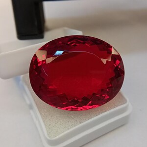 May include: A large, faceted, oval-shaped, deep red gemstone. The gem is displayed in a white box with a foam insert. The gem's surface reflects light, showcasing its intricate cuts and vibrant color. The background is a neutral tone.