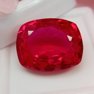 Può includere: Una gemma dal taglio a cuscino con un colore rosso intenso e saturo, caratterizzata da una superficie sfaccettata che riflette la luce. La pietra è posizionata su un contenitore bianco quadrato con un dettaglio in tessuto rosa chiaro.