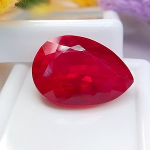 Könnte beinhalten: Ein birnenförmiger, facettierter Edelstein in einer tiefen, leuchtend roten Farbe. Der Stein hat eine polierte Oberfläche mit mehreren geometrischen Facetten, die das Licht reflektieren und für ein brillantes Aussehen sorgen. Er liegt auf einer glatten, weißen Oberfläche.