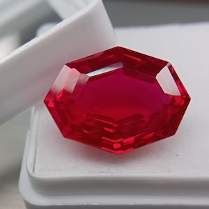 Könnte beinhalten: Ein leuchtend tiefroter Edelstein mit einem facettierten, länglichen Hexagonschliff. Der Stein weist eine hohe Klarheit und interne Lichtreflexion auf und liegt auf einem weißen Präsentationssockel mit einem teilweise sichtbaren weißen Deckel.