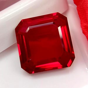 Könnte beinhalten: Ein quadratisch geschliffener, leuchtend roter Edelstein mit facettierten Kanten, die scharfe geometrische Reflexionen erzeugen. Der Stein zeigt einen tiefen, gesättigten Karmesinton und eine hochtransparente Oberfläche.