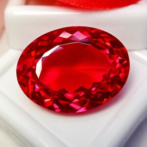 May include: A vibrant, oval-cut gemstone with a deep red color and multifaceted surface that reflects light. The stone is positioned on a white display base, showcasing its transparent clarity and polished, symmetrical edges throughout.