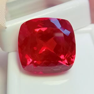 May include: A vibrant, cushion-cut red gemstone with a faceted surface that reflects light, creating deep crimson highlights and shadows. The gemstone is positioned against a smooth, white, rectangular display base with rounded corners.