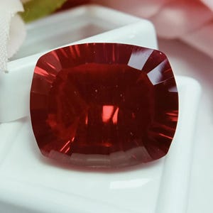 Peut inclure: Une pierre précieuse rouge foncé de taille coussin avec une surface à facettes créant un motif radial de reflets lumineux. La pierre présente une forme rectangulaire aux coins arrondis et un aspect transparent et poli, révélant une teinte cramoisie.