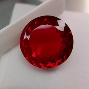 Könnte beinhalten: Ein facettierter, runder, tiefroter Edelstein. Der Edelstein ist hochglanzpoliert, reflektiert das Licht und erzeugt ein brillantes Aussehen. Der Edelstein ist vor einem weißen Hintergrund platziert, der seine Farbe und Klarheit hervorhebt. Geeignet für die Schmuckherstellung.