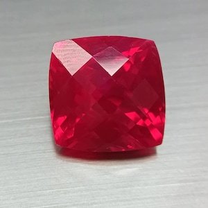 May include: A faceted, cushion-cut gemstone in a vibrant shade of red. The gem's surface is a complex arrangement of angled planes, reflecting light and creating a sparkling effect. The gemstone is set against a neutral background.