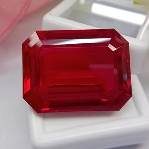 May include: A vibrant, deep red gemstone with an emerald-cut shape, featuring sharp, clean facets that reflect light across its surface. The rectangular stone is positioned directly on top of a small, white, rectangular plastic display case.