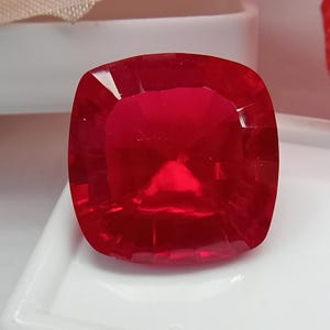 Pode incluir: Uma gema vermelha vibrante de corte almofada com uma superfície facetada que reflete a luz. A pedra tem um formato quadrado com cantos arredondados e uma cor carmesim translúcida profunda, exibindo cortes geométricos precisos na superfície.