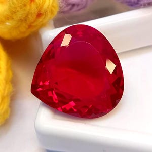 Peut inclure: Une pierre précieuse rouge vif et profonde taillée en forme triangulaire avec de multiples facettes qui reflètent la lumière. La pierre présente une surface polie et une teinte cramoisie saturée et brillante, avec une transparence claire.